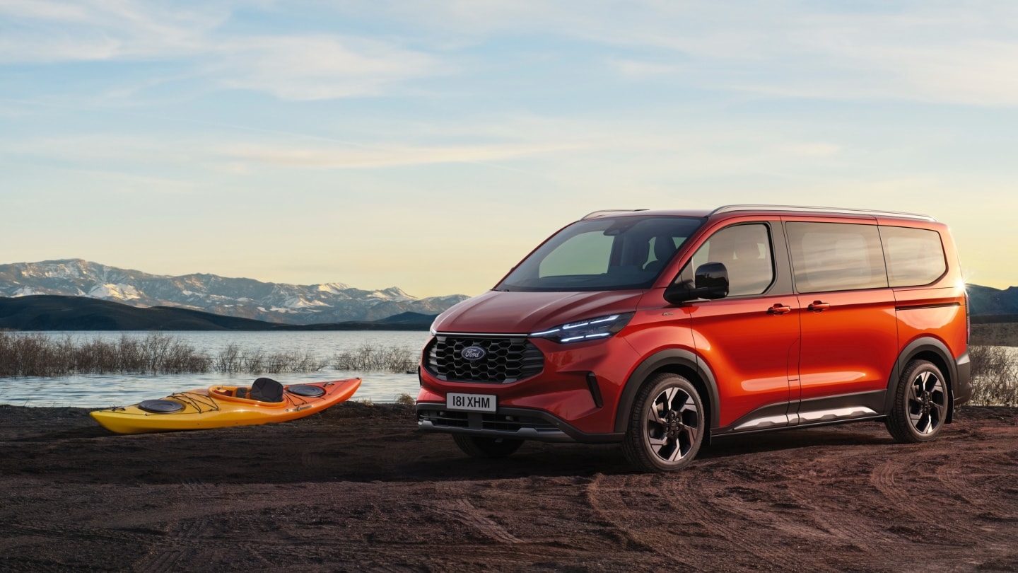 A red Ford Tourneo Connect parked by a lake, with a kayak and snow-capped mountains in the background.