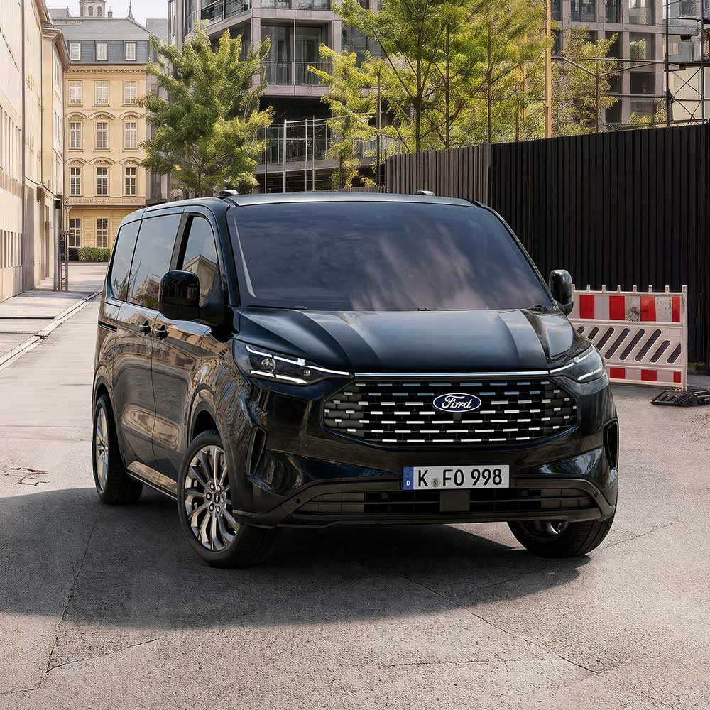 Ein schwarzer Ford Tourneo Custom steht auf einer Straße vor Gebäuden, mit einer Baustellenabsperrung rechts. 