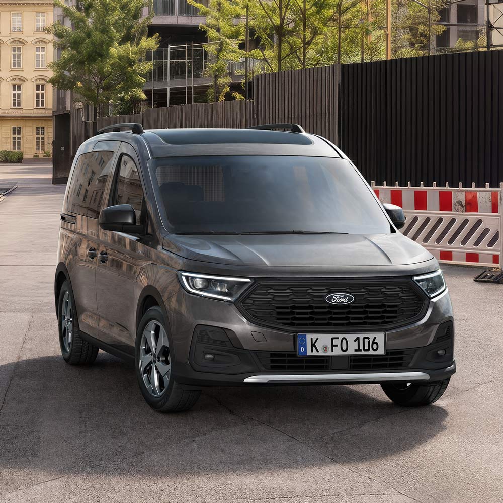 Ein dunkelgrauer Ford Tourneo Connect steht auf einer Straße vor Gebäuden, mit einer Baustellenabsperrung rechts. 