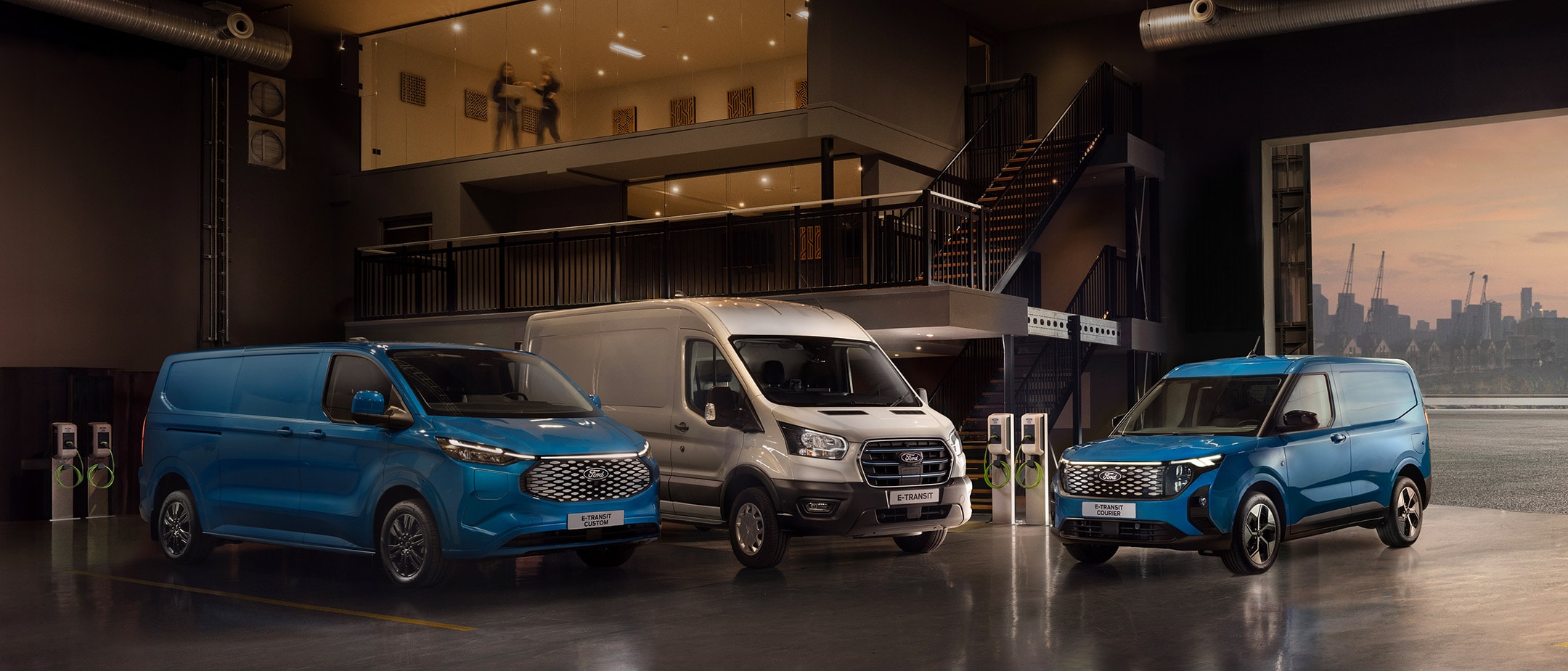 Drei elektrische Ford-Transporter (E-Transit Custom, E-Transit, E-Transit Courier) parken in Innenräumen mit Ladestationen.	