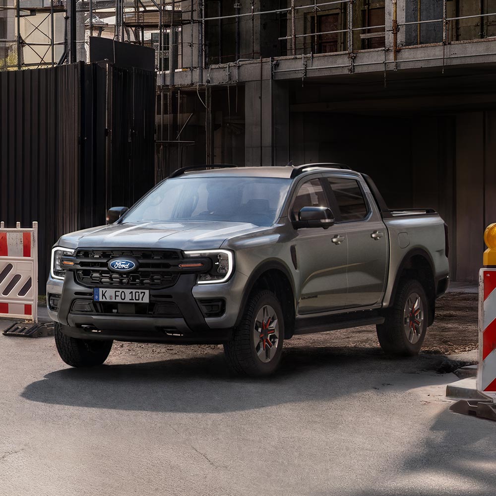 Ein grauer Ford Ranger steht auf einer Straße vor Gebäuden, mit einer Baustellenabsperrung links. 