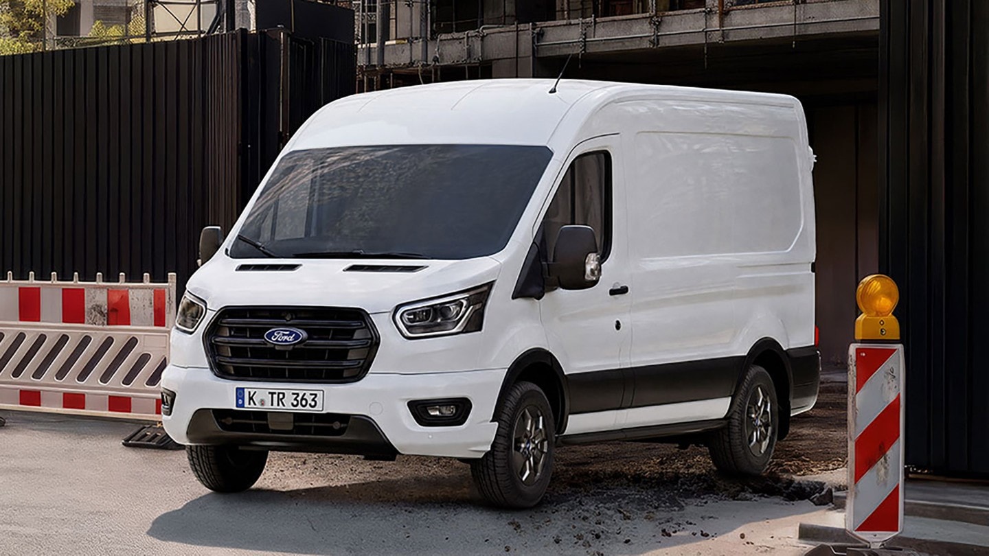 Ein weißer Ford Transit Kastenwagen steht auf einer Straße neben Baustellenabsperrungen und einer Warnleuchte. 