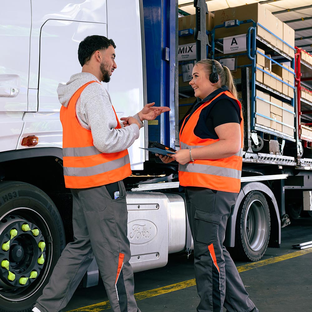 Zwei Logistikfachkräfte in orangefarbenen Warnwesten sprechen vor einem Lkw.
