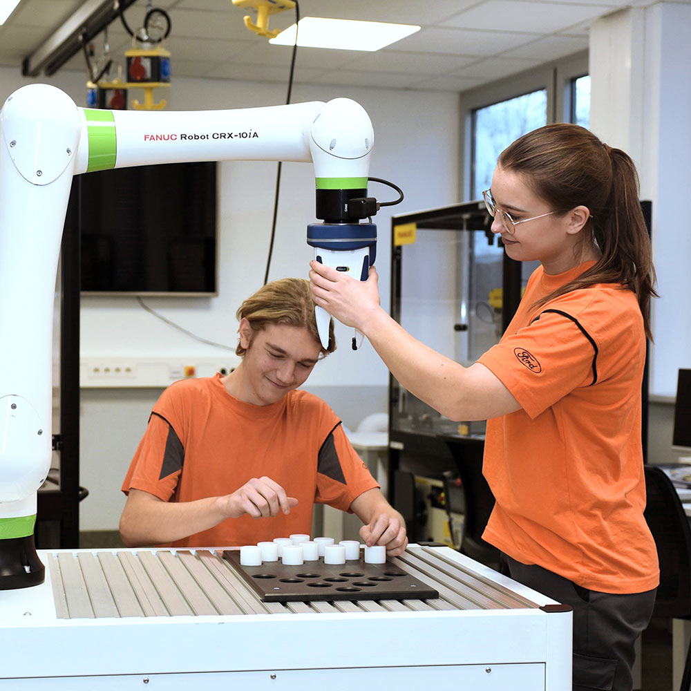 Zwei Personen bedienen einen weißen FANUC-Roboterarm in einer Werkstatt.