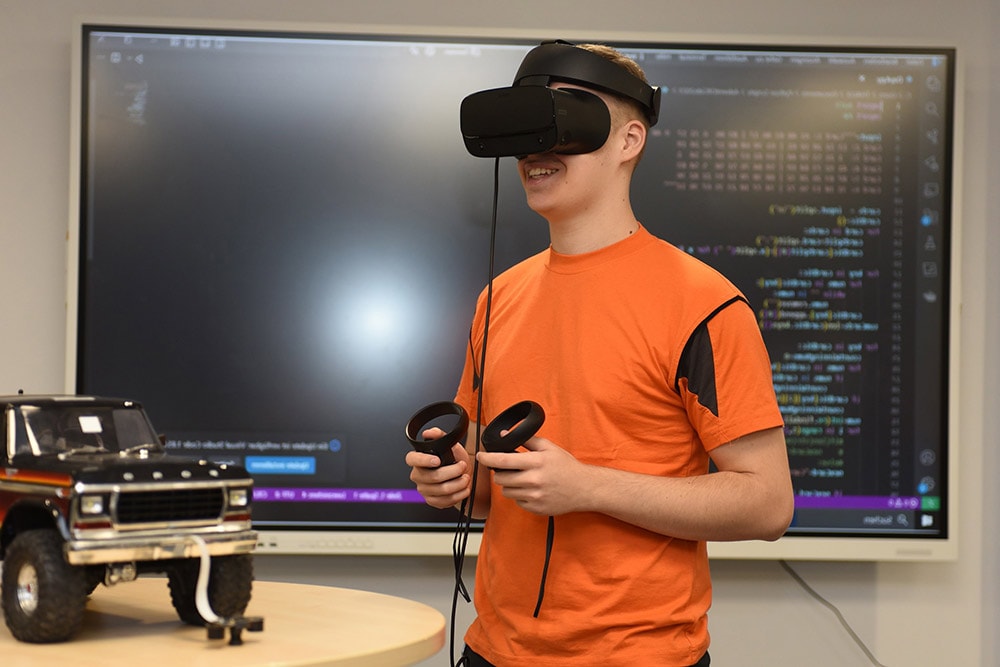 Eine Person trägt ein VR-Headset und hält Controller vor einer Anzeigetafel.