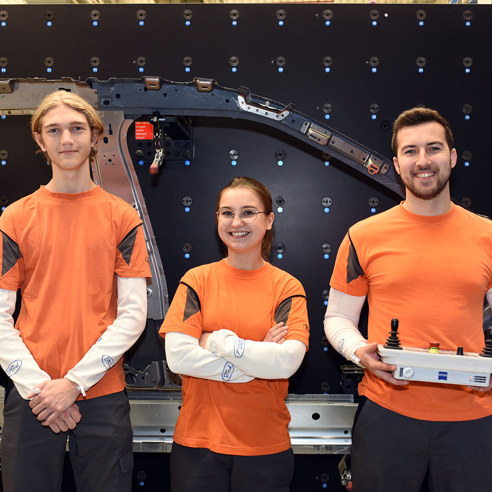 Drei Personen in orangefarbenen Ford-Arbeitsshirts stehen vor einer schwarzen Messwand.