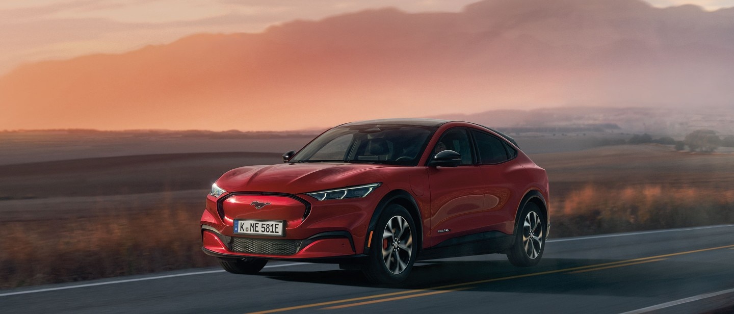 Ford Mustang Mach-E ¾-Frontansicht in Red, am Straßenrand stehend vor einer Wüstenlandschaft, ein Mann und eine Frau gehen auf der Straße
