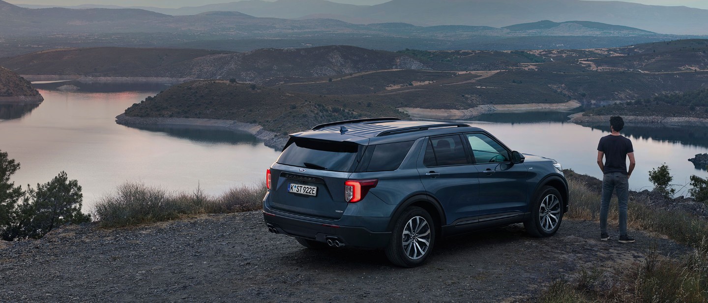 Ford Explorer Plug-in-Hybrid in Blau. ¾-Heckansicht. Ein Mann steht neben dem Fahrzeug vor einer Landschaft mit Bergen und Seen