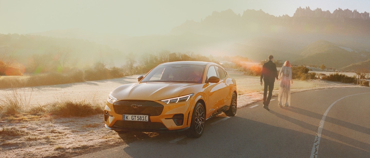 Ford Mustang Mach-E ¾-Frontansicht in Orange, am Straßenrand stehend vor einer Wüstenlandschaft, ein Mann und eine Frau gehen auf der Straße