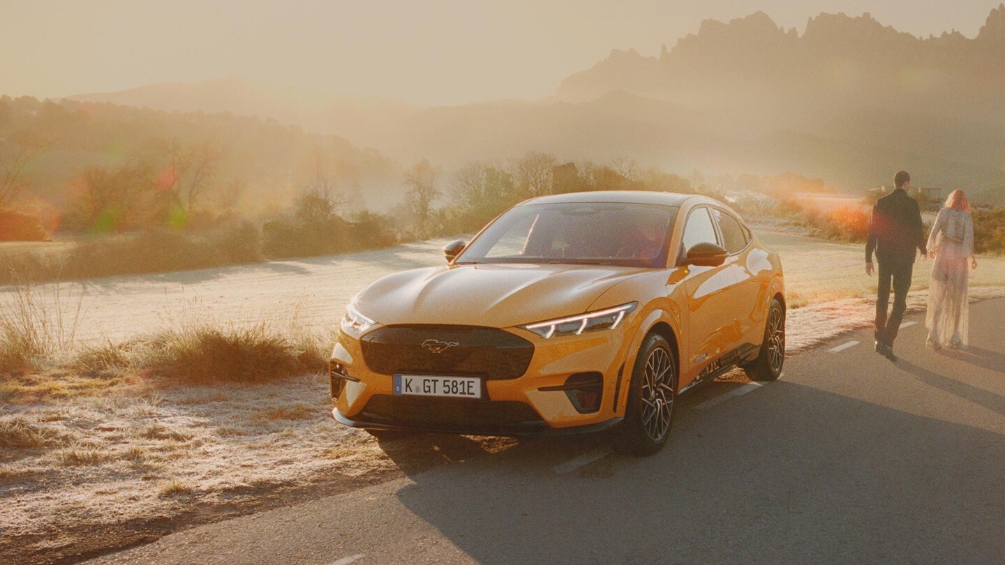 Ford Mustang Mach-E GT in Orange. Dreiviertel-Frontansicht, auf einer Straße vor Bergen im Sonnenuntergang fahrend.