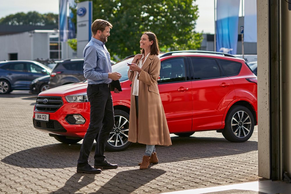 Ein Mann und eine Frau unterhalten sich neben einem roten Ford Kuga auf einem Händlerparkplatz