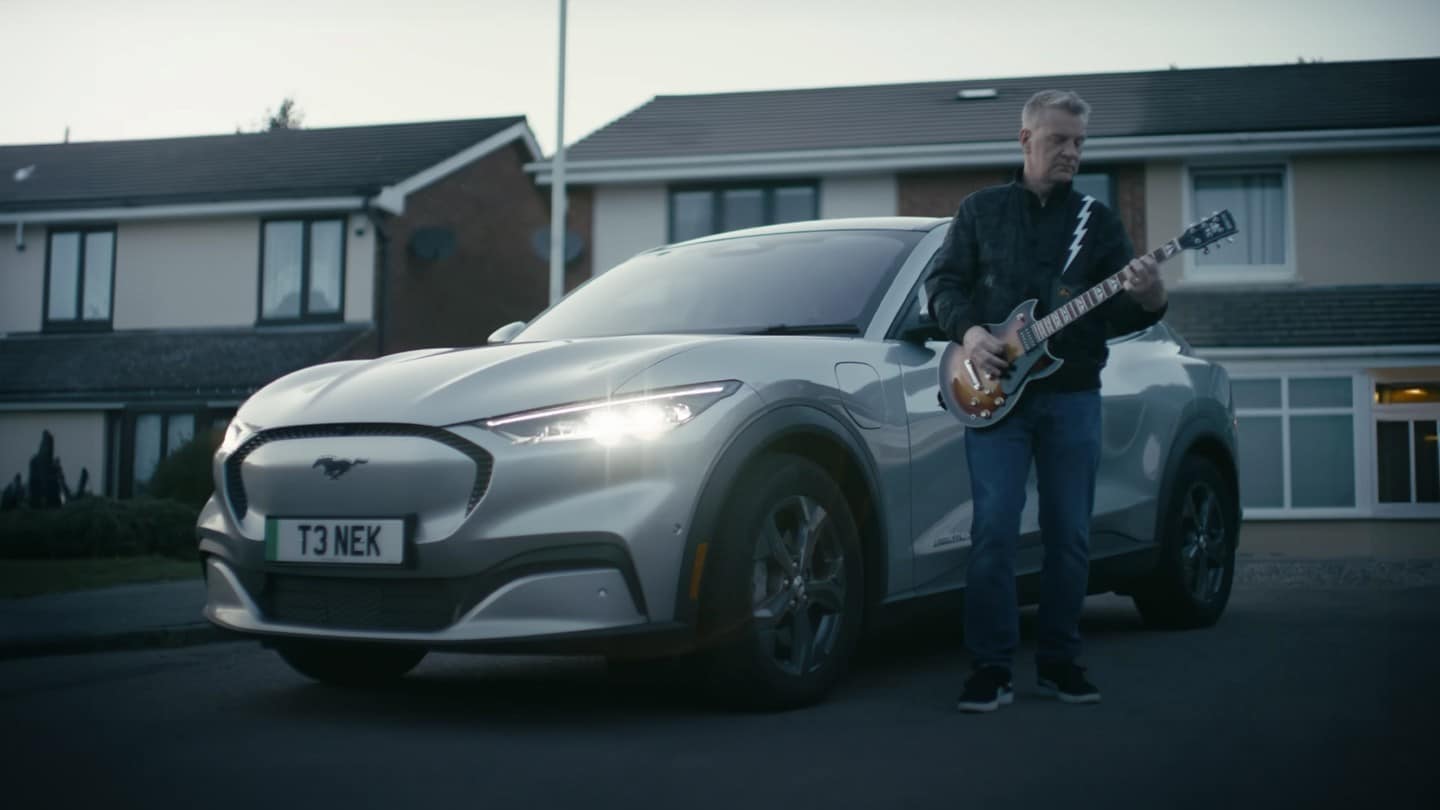 Der Musiker Peter Steer steht mit seiner Gitarre neben seinem Mustang Mach-E.