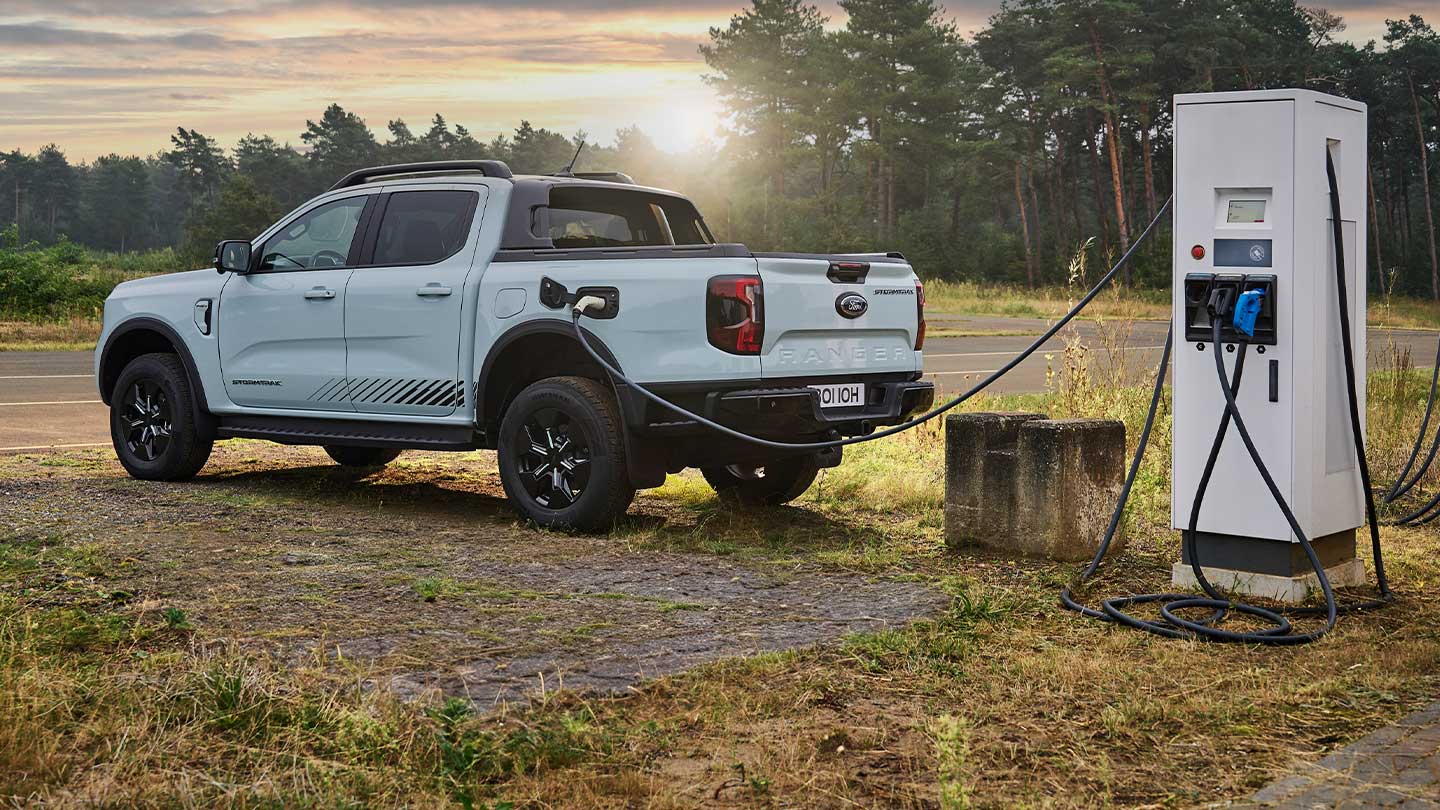 Ein weißer Ford Ranger Plug-in-Hybrid steht an einer Ladestation im Freien und wird aufgeladen. Im Hintergrund geht die Sonne in einer bewaldeten Landschaft auf.
