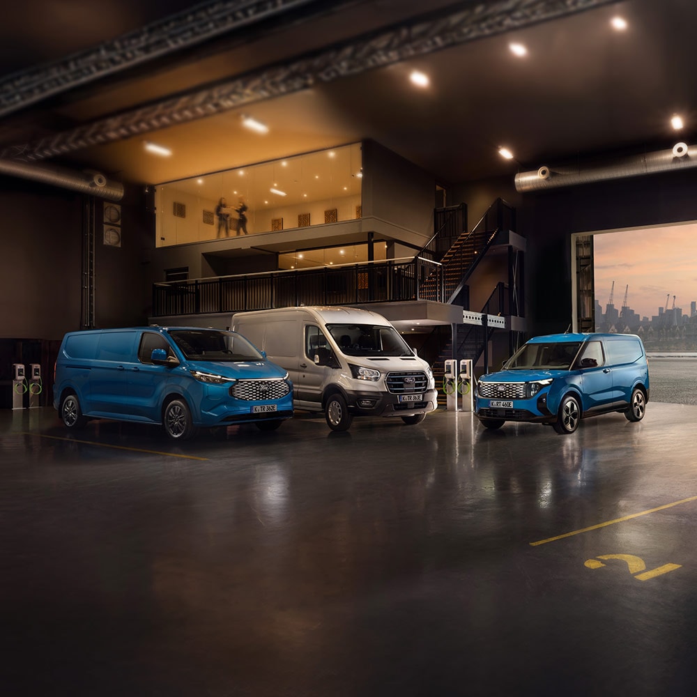Drei elektrische Ford-Nutzfahrzeuge in einer Ladehalle mit Ladestationen im Hintergrund.