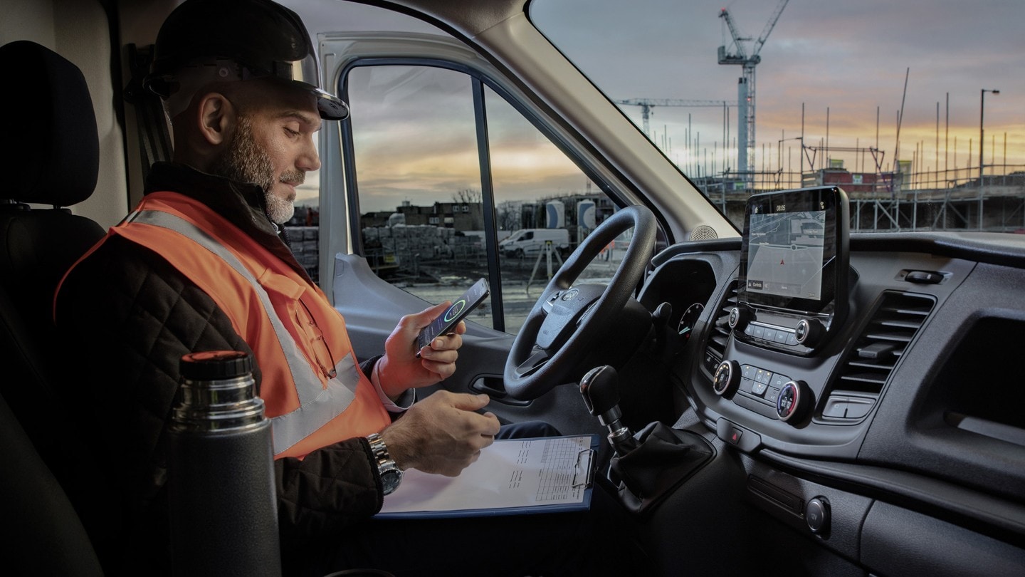 Arbeiter mit Bauhelm sitzt hinter dem Steuer eines Fahrzeugs und schaut auf ein mobiles Telefon