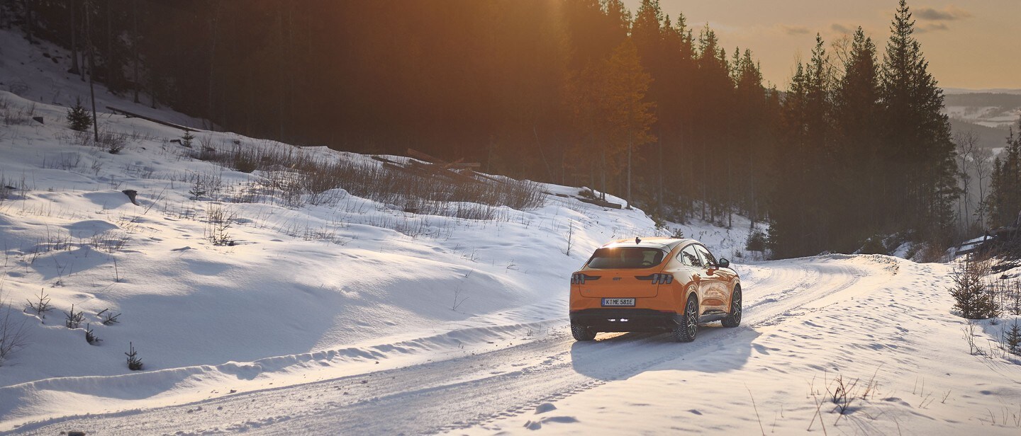Ford Mustang Mach-E in Orange fahrend auf Straße im Schnee in Berglandschaft