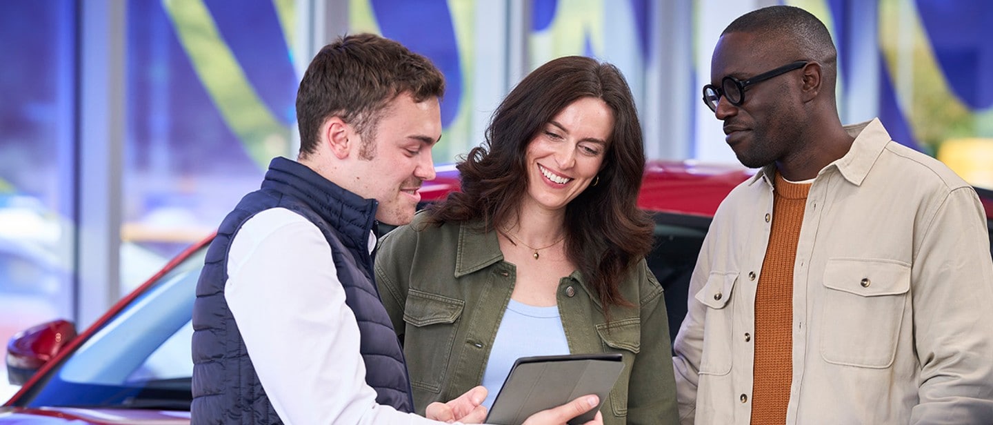 Zwei Männer und eine Frau stehen zusammen in einem Autohaus, lächeln und schauen auf ein Tablet, das einer der Männer hält. Im Hintergrund ist ein rotes Auto zu sehen.