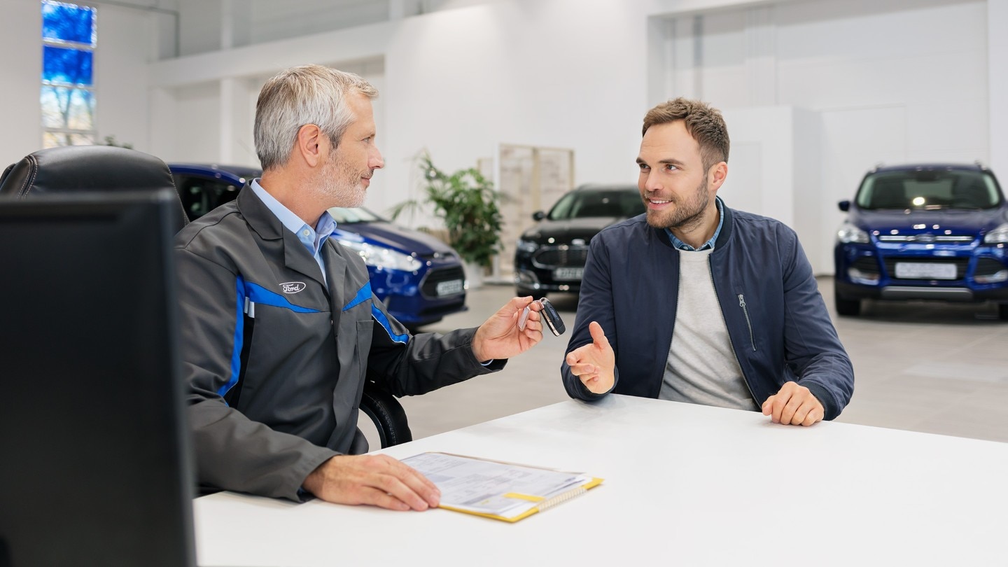 Ein Ford-Mitarbeiter übergibt Autoschlüssel an einen Kunden an einem Schreibtisch, mit Ford-Fahrzeugen im Hintergrund.