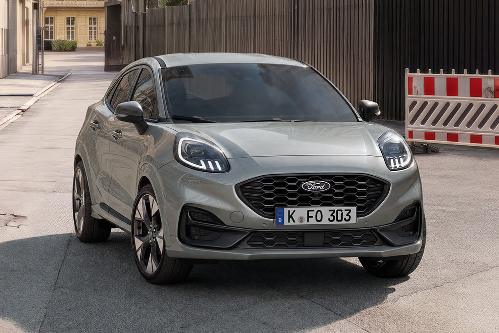 Ein grauer Ford Puma steht auf einer Straße, von vorne schräg gesehen, mit Absperrungen im Hintergrund.
