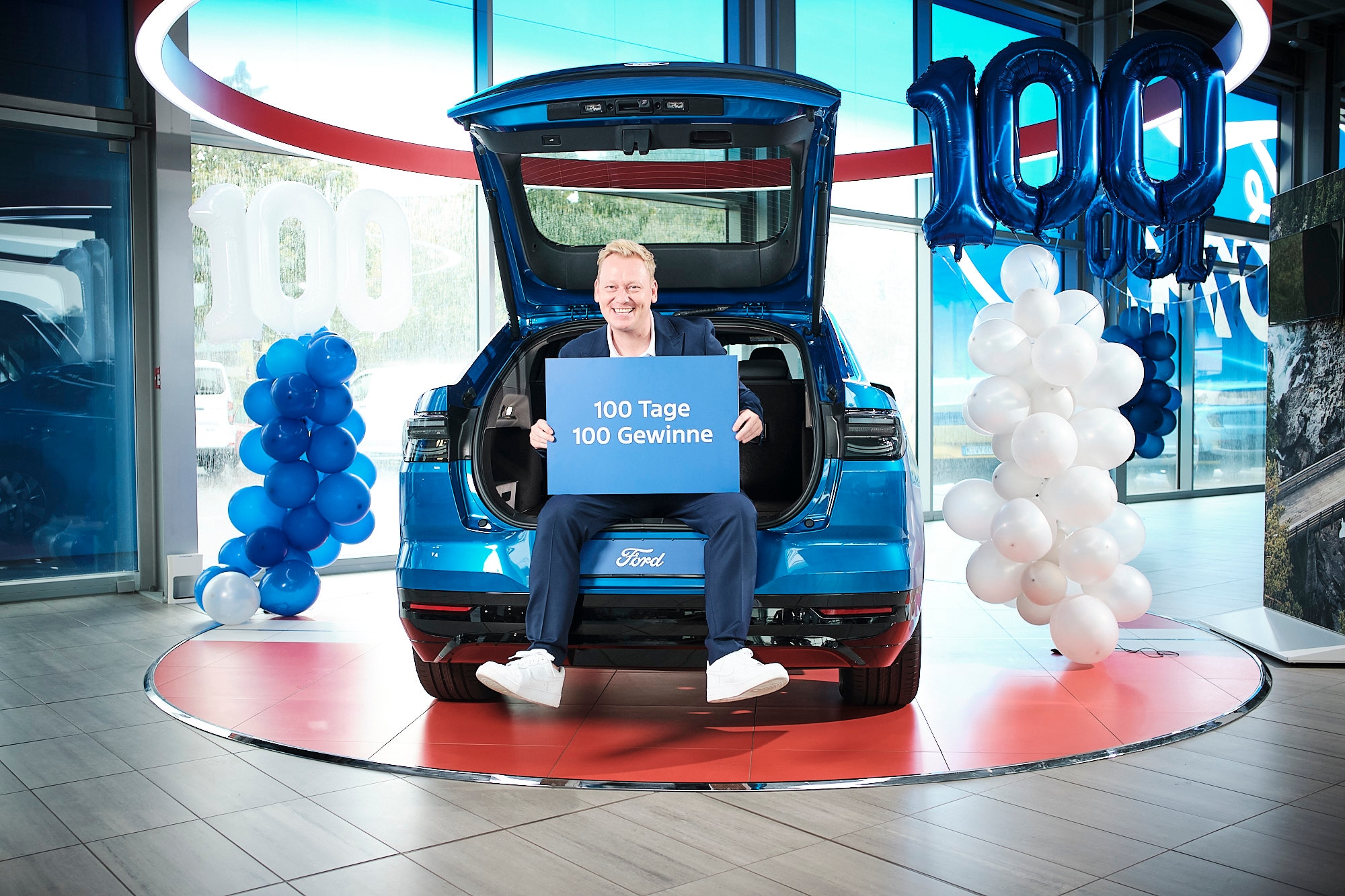 Der deutsche Entertainer „Knossi“ sitzt zwischen Ballons im Kofferraum des Ford Capri mit einem Schild „100 Tage 100 Gewinne“