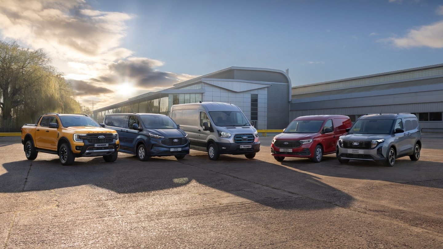 Vier Ford-Nutzfahrzeuge (E-Transit Custom, E-Transit, E-Transit Courier und Transit Connect PHEV) vor dem Hintergrund eines Innenraums mit Treppe und einem Fenster mit Blick auf eine Stadtlandschaft. Die Vans sind in verschiedenen Farben gehalten, darunter Blau und Grau.