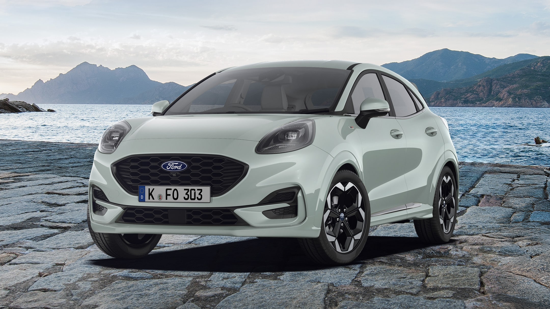 Ein grauer Ford Puma, parkt auf Pflastersteinen. Im Hintergrund erstreckt sich eine Berglandschaft mit Wasser.