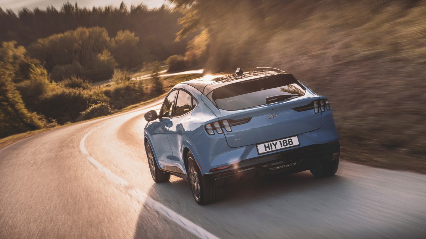 Ford Mustang Mach-E in Blau 3/4-Heckansicht auf einer Landstraße fahrend