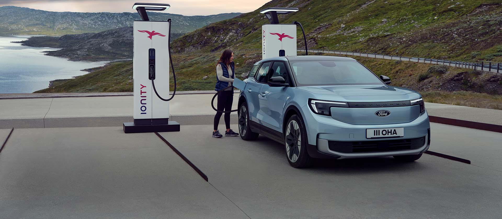 Eine Frau schließt einen blauen, vollelektrischen Ford Explorer mit einem Kabel an eine öffentliche Ladestation an. Das Auto ist in der Nähe von Bergen und Wasser geparkt.