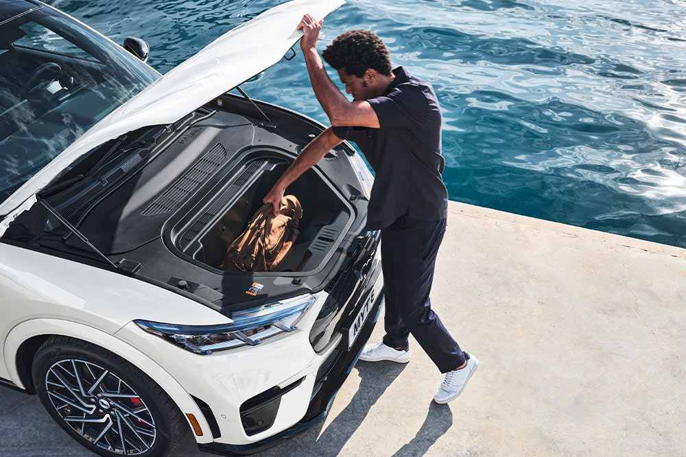 Eine Person legt eine Reisetasche in den Kofferraum eines weißen Ford Mustang Mach-E Elektrofahrzeugs. 