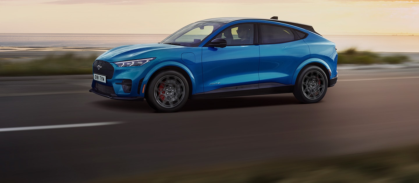 Ein blauer elektrischer Ford Mustang Mach-E fährt bei Sonnenuntergang eine Küstenstraße entlang.