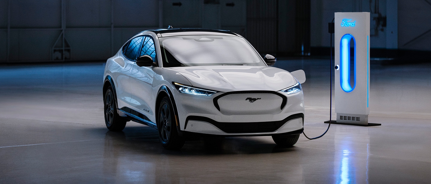 Ein weißer Ford Mustang Mach-E steht in einer Garage und ist an eine blau leuchtende Ladestation angeschlossen.