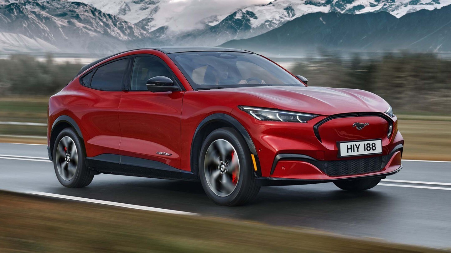 Ford Mustang Mach-E in Rot 3/4-Frontansicht fahrend auf einer Landstraße mit Bergen im Hintergrund