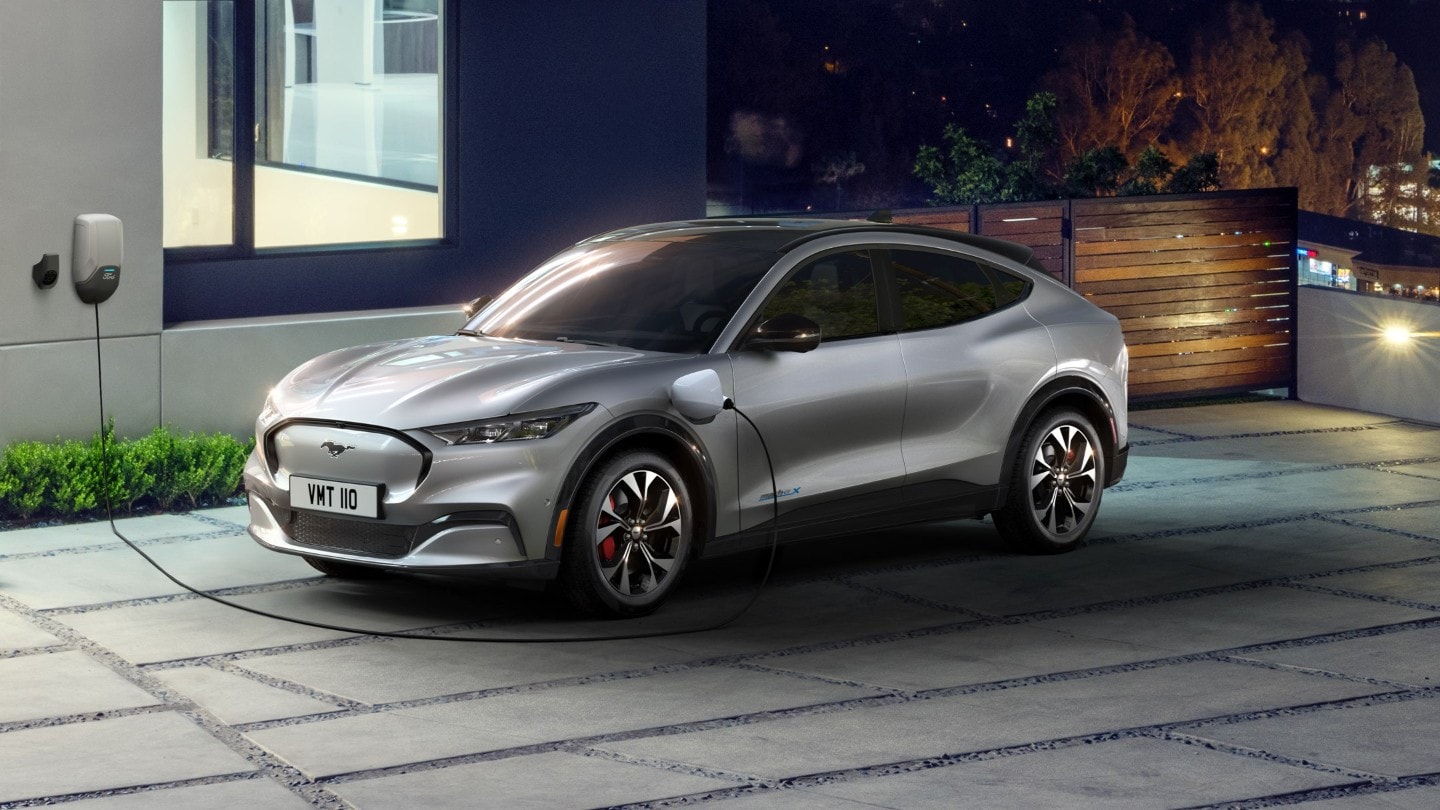 Ford Mustang Mach-E ¾-Frontansicht in Grau parkend vor Wohnhaus beim Laden über eine Ladestation