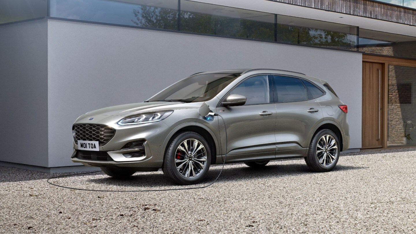 Ford Kuga in Grau ¾-Frontansicht parkend vor Wohnhaus beim Laden über eine Steckdose