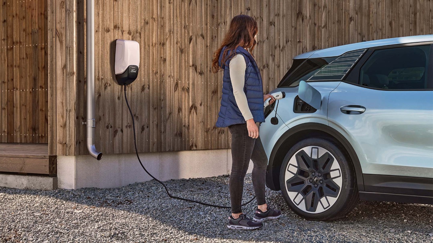 Der neue vollelektrische Ford Explorer an einer Wallbox parkend, eine Frau nebenstehend hält ein Ladekabel