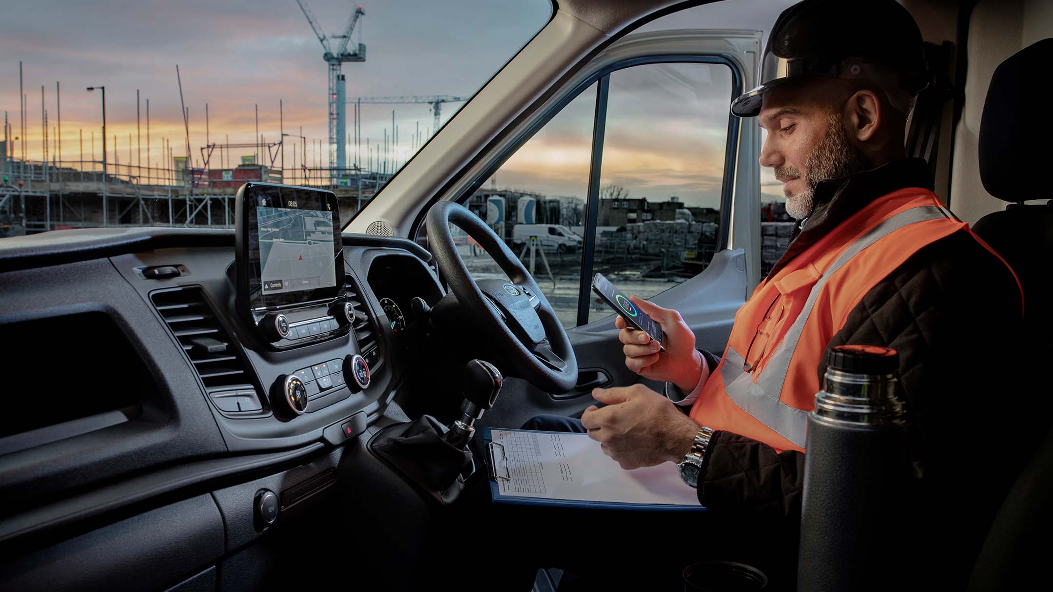 Arbeiter mit Bauhelm sitzt hinter dem Steuer eines Fahrzeugs und schaut auf ein mobiles Telefon