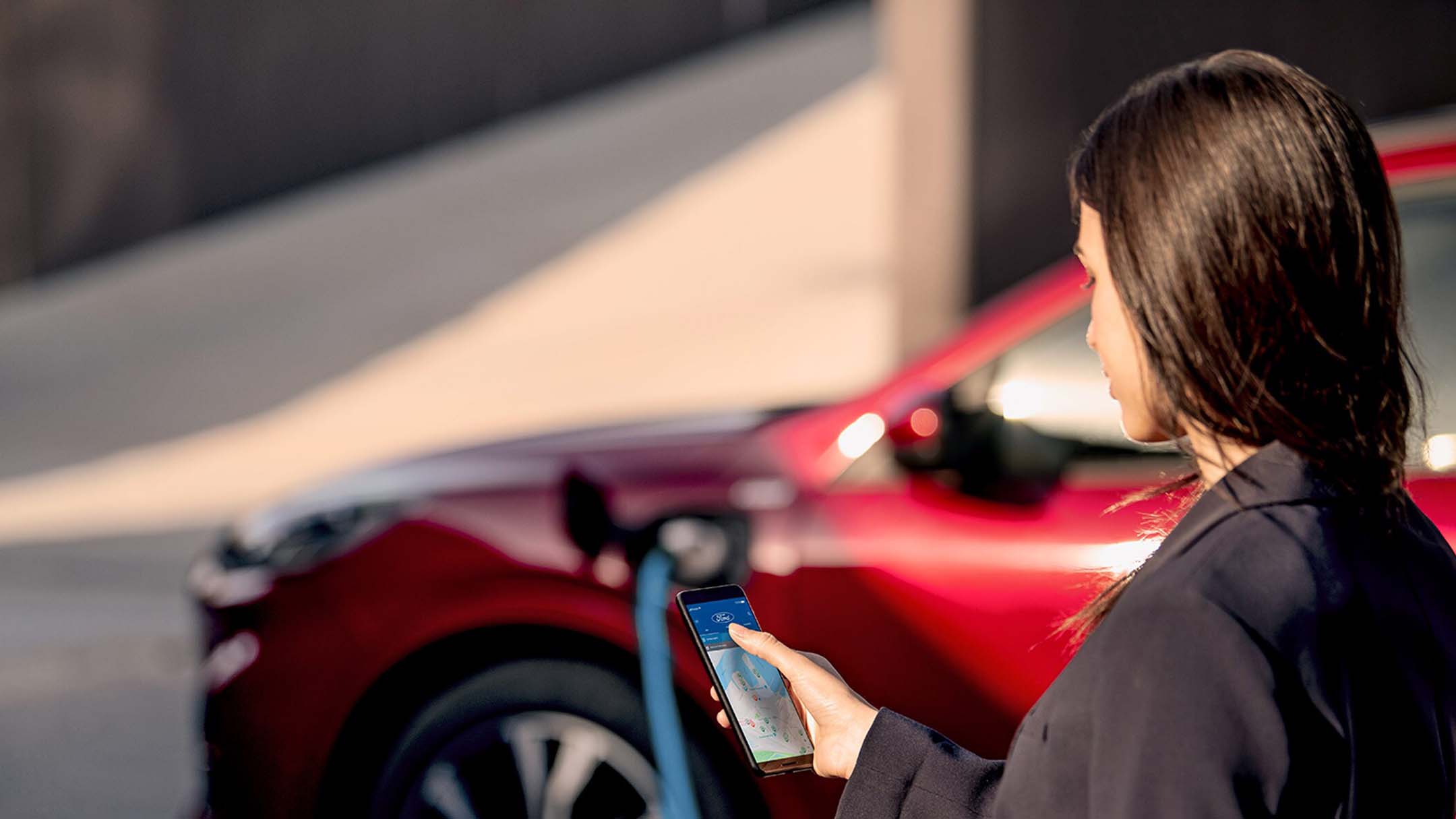 Frau mit Smartphone beim elektrischen Laden ihres Ford Fahrzeugs