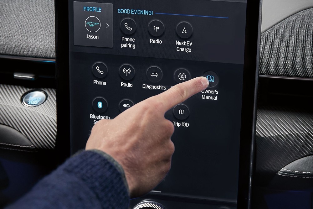 Eine Hand tippt auf den Touchscreen eines Ford-Infotainmentsystems, das ein Menü mit Optionen wie "Phone", "Radio" und "Owner's Manual" anzeigt.