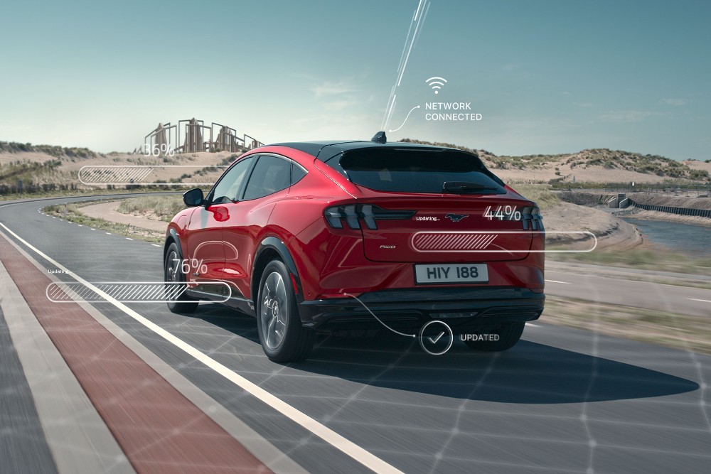 Ein roter Ford Mustang Mach-E fährt auf einer Straße, von hinten rechts gesehen, mit Grafiken, die drahtlose Konnektivität und Datentransfer andeuten. Im Hintergrund ist eine weite, offene Landschaft zu sehen.