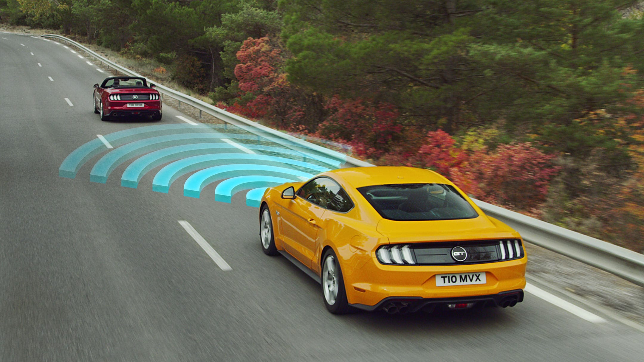 Ford Mustang in Orange. Heckansicht, fahrend über eine Landstraße hinter einem roten Auto mit visualisierter Geschwindigkeitsregelanlage