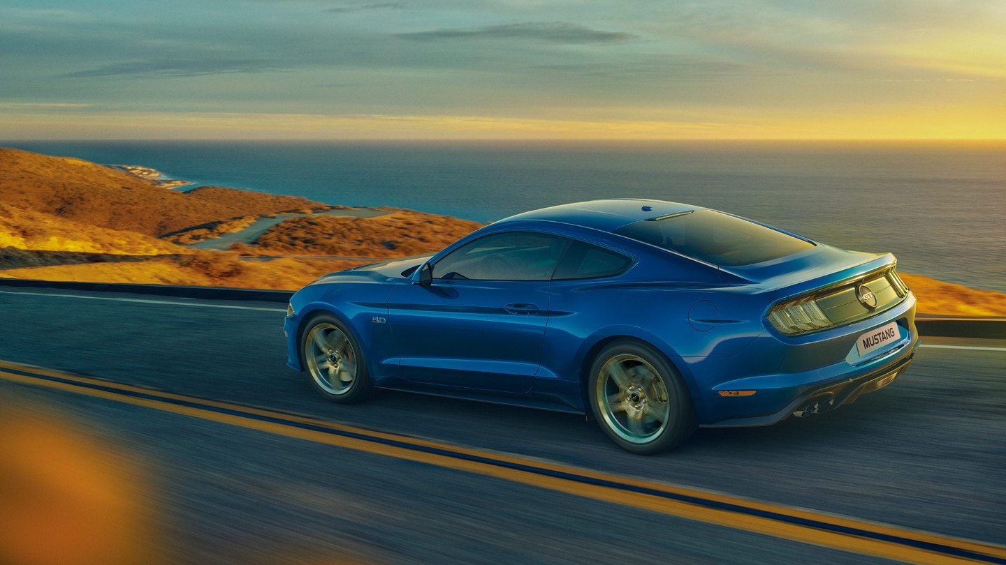 Ford Mustang in Blau. Seitenansicht, fahrend auf einer Landstraße an einem Meer mit Sonnenaufgang im Hintergrund