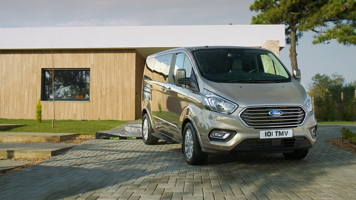 Ford Tourneo Custom Champagner ¾-Frontansicht parkt vor Holzhaus