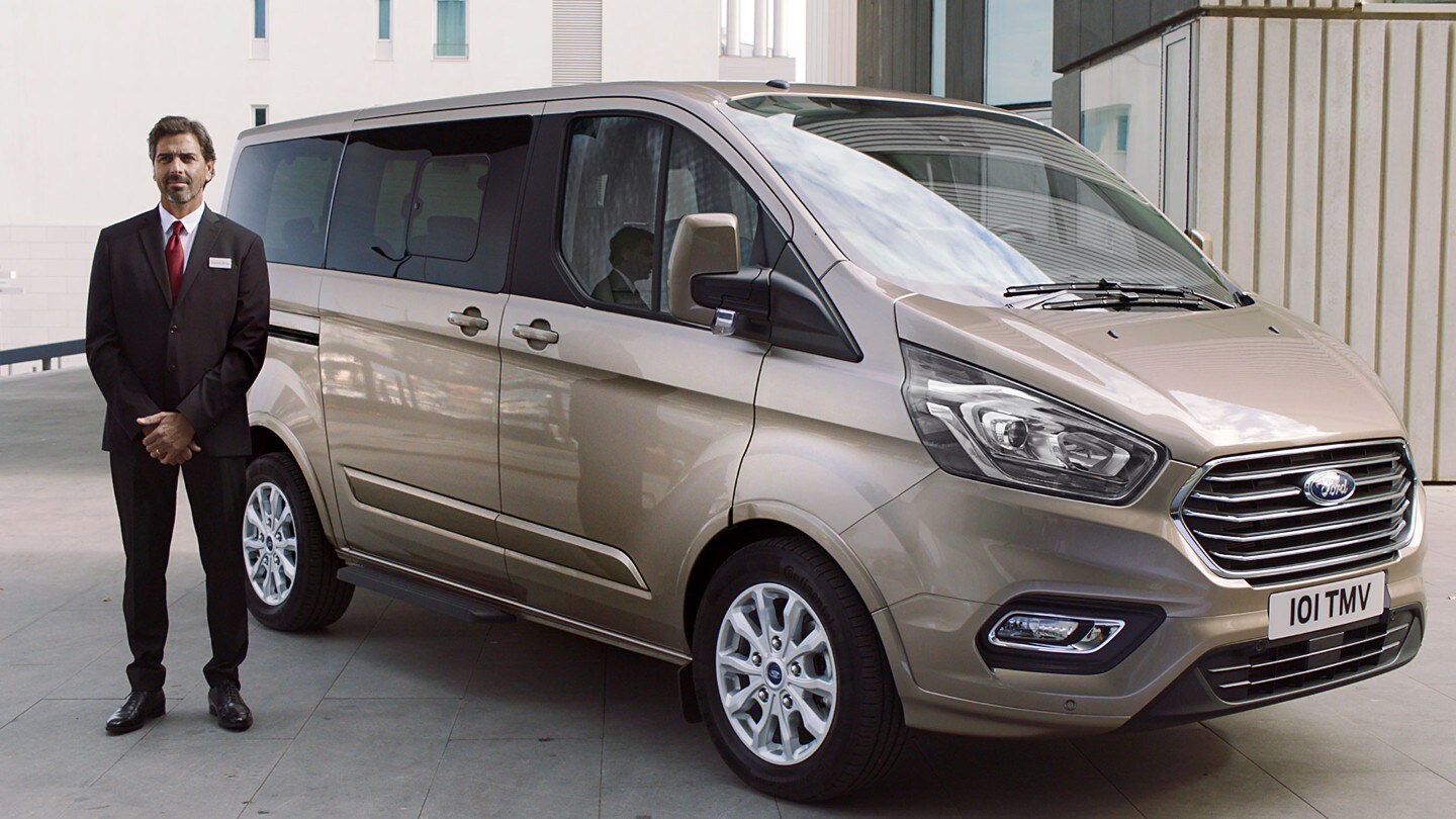Ford Tourneo Custom Champagner ¾-Seitenansicht parkt neben modernem Gebäude Mann steht auf Beifahrerseite