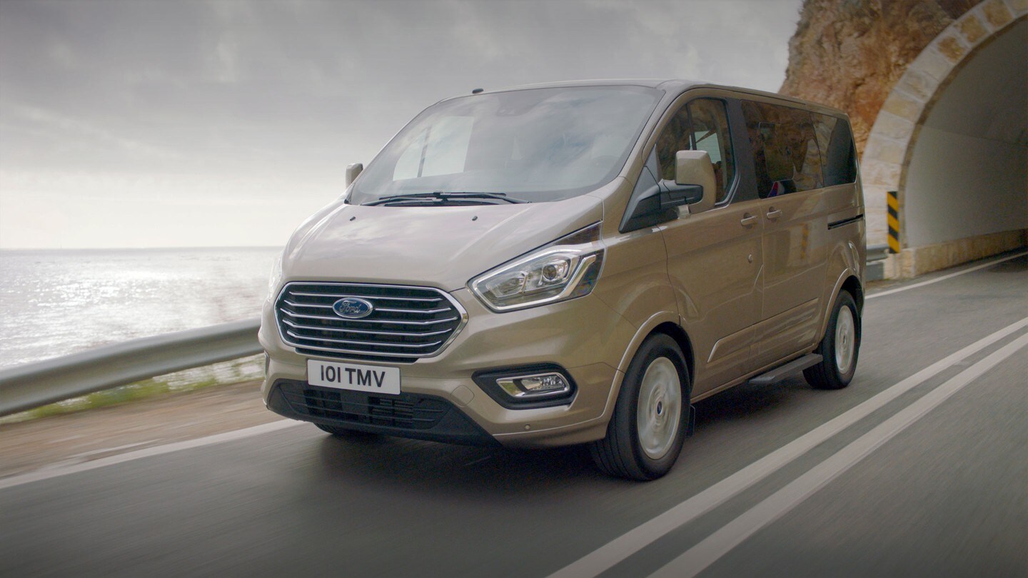 Ford Tourneo  Custom Champagner ¾-Frontansicht in Bewegung auf Landstraße hinter Tunnel 