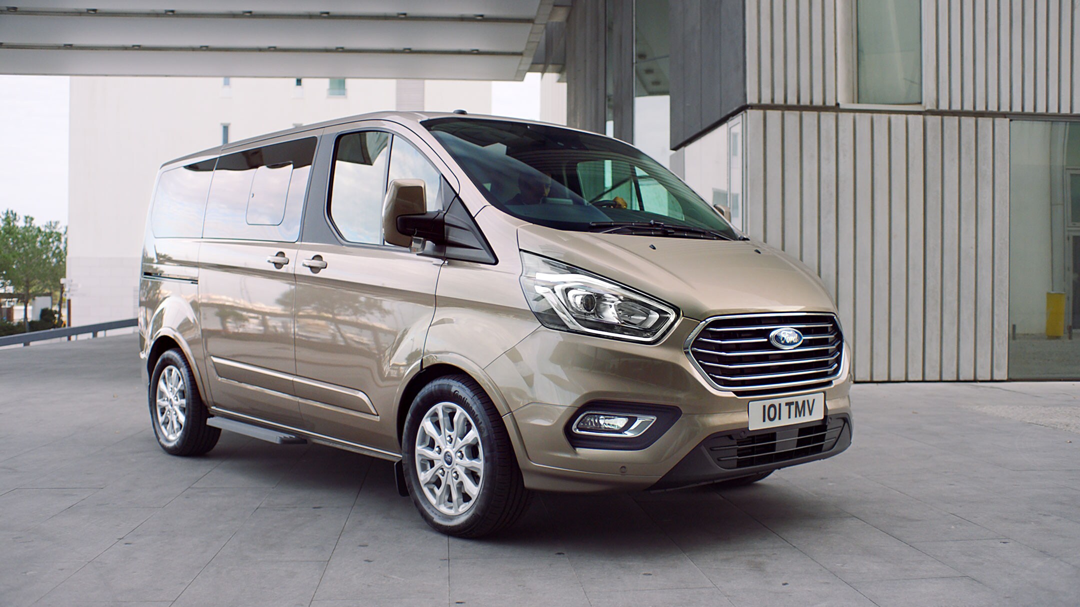 Ford Tourneo  Custom Champagner ¾-Frontansicht parkt vor modernem Gebäude