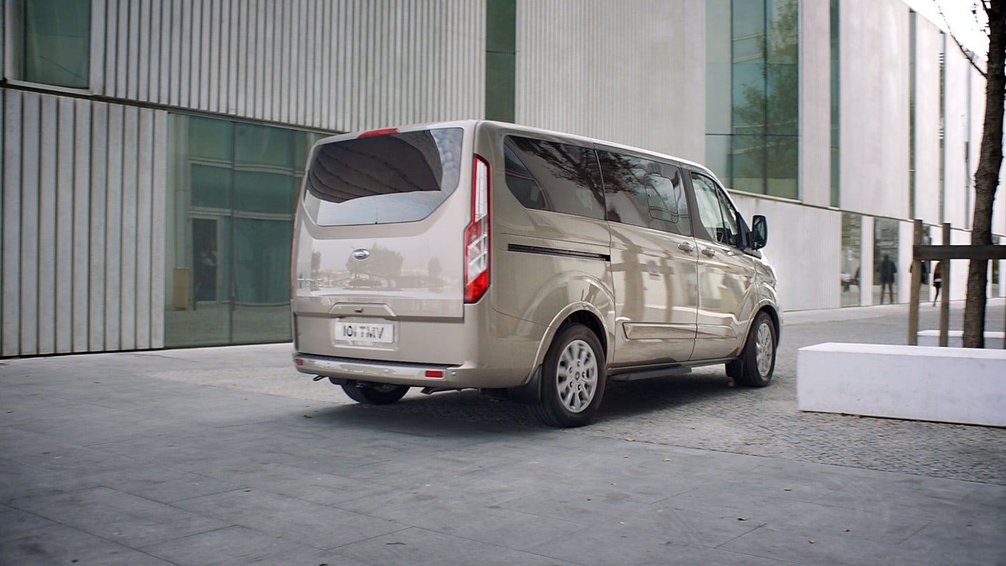 Ford Tourneo  Custom Silber ¾-Heckansicht parkt vor modernem Gebäude