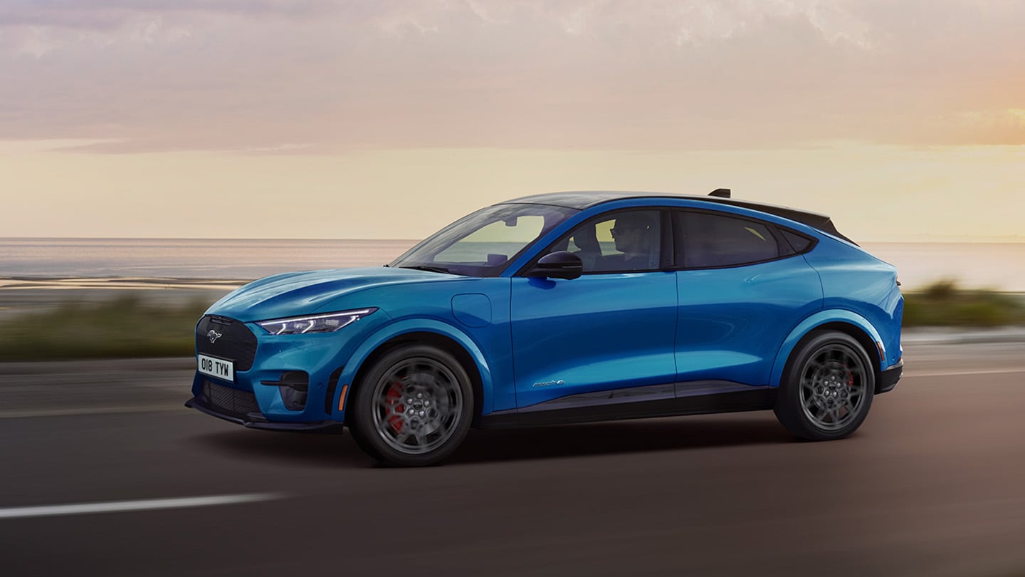 Seitenansicht eines blauen elektrischen Ford Mustang Mach-E, der in der Abenddämmerung eine Küstenstraße entlangfährt.