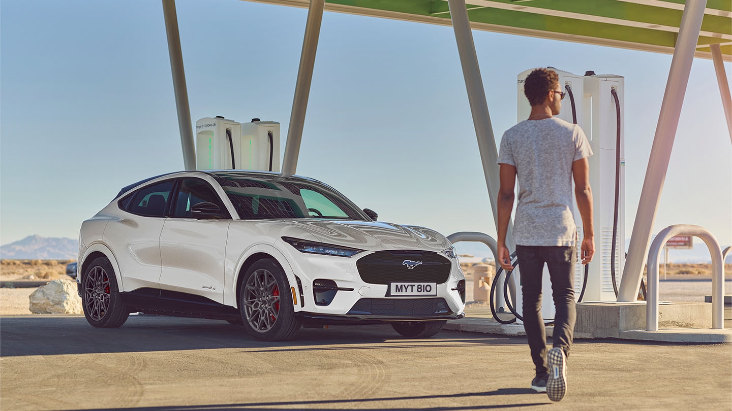 Ein weißer Ford Mustang Mach-E, der an einer öffentlichen Ladestation parkt. Ein Mann in einem weißen T-Shirt geht auf das Auto zu.