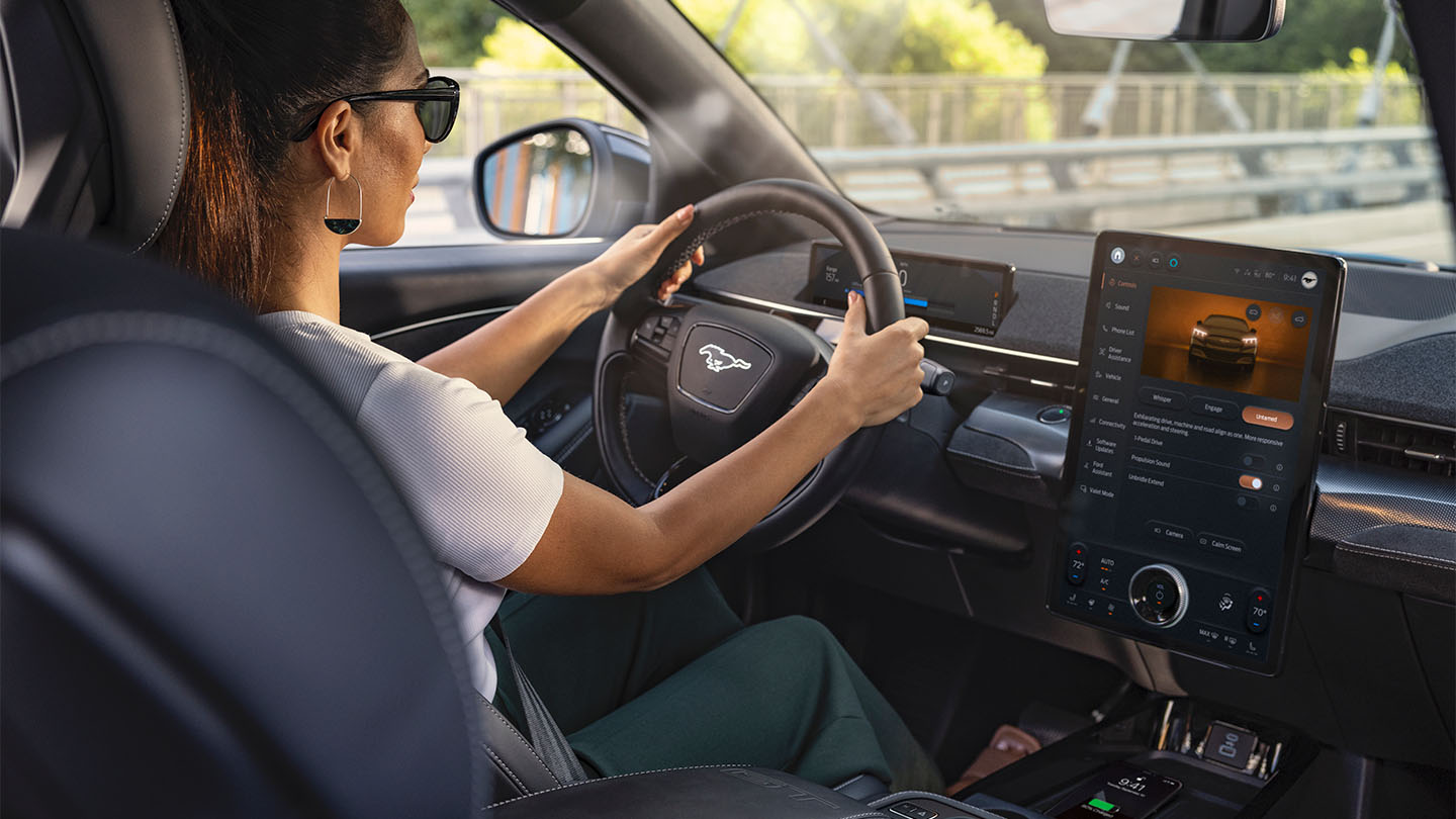 Eine Frau, die den Mustang Mach-E fährt und dabei die Hände am Lenkrad hat. Der 15,5-Zoll-Touchscreen zeigt Details zum Fahrzeug an.