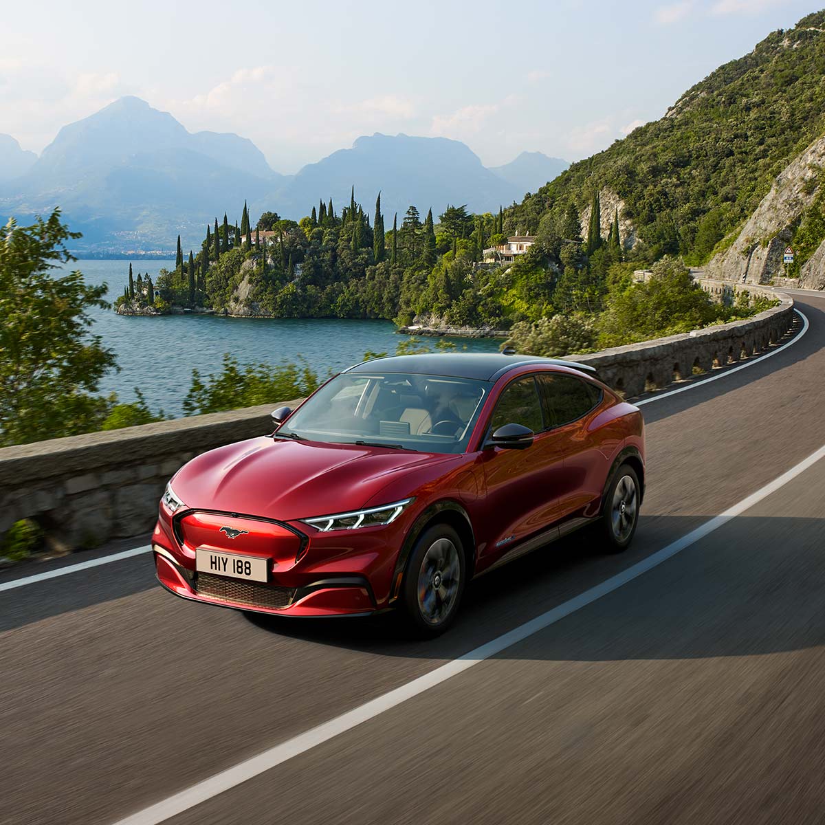 Ford Mustang Mach-E in rot. Dreiviertelansicht, fahrend auf Bergstraße vor See